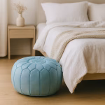A Moroccan leather pouf blue placed beside a minimalist wooden bed in a cozy, light-filled bedroom.