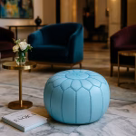 A Moroccan leather pouf blue styled in a chic hotel lobby with velvet armchairs and gold accents.