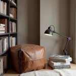 Square leather ottoman pouf in reading nook corner beside bookshelf with task lamp and books
