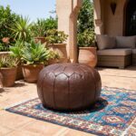 An outdoor patio area with a Chocolate Brown Ottoman resting on a blue patterned rug, surrounded by terracotta pots and succulent plants.