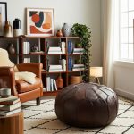 Dark Chocolate Brown Ottoman on a geometric rug near a cognac leather armchair and a wooden bookshelf with plants.