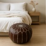 Dark brown leather round pouf with geometric stitching beside a wooden bed in a minimalist bedroom