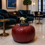 Deep Red Leather Pouf Round styled in a boutique lounge with teal armchairs and gold tables.