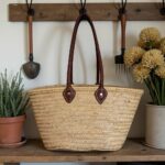 Handwoven straw tote bag with leather handles hanging on wooden hooks - sustainable market basket