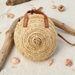 Round straw bag styled on beach with seashells, handwoven circle basket with leather straps for vacation