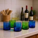 Five mixed-tone blue and green moroccan wine glasses displayed on a kitchen counter with wine bottles behind.