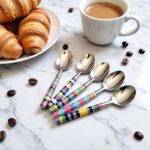 Set of 5 ceramic tea spoons with rainbow striped handles beside croissants and coffee on marble countertop