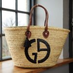Designer straw tote bag by window showing black logo and brown leather handles