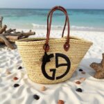 Designer straw tote bag with black monogram on sandy beach with seashells