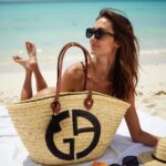 Woman at beach with designer straw tote bag featuring black emblem and leather straps