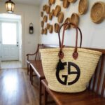 Designer straw tote bag with black logo detail displayed on wooden bench in home setting