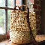 Close-up of straw backpack showing chocolate leather straps and open lattice weave by brick window