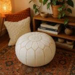 White leather round ottoman floor pouf with hand-stitched detail on Persian rug beside bookshelf and pillows