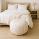 White leather round ottoman floor pouf at foot of bed in minimalist bedroom with wooden furniture