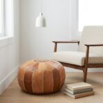 Minimalist home decor featuring a Leather Round Ottoman next to a mid-century modern armchair and a stack of books under a white pendant light.