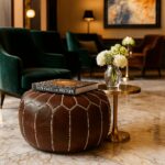 Dark brown leather round pouf with traditional stitching in a sophisticated hotel lounge beside velvet armchairs
