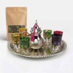 A vibrant hand-hammered silver beldi tea set featuring a teapot accented with a pink tassel and six colorful, patterned tea glasses arranged on a large engraved silver tray, displayed next to a kraft paper pouch of loose leaf green tea against a white background.
