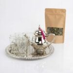 A complete silver tea gift set with engraved teapot featuring a bright pink tassel, accompanied by six clear tea glasses with intricate silver metalwork bases. The collection is arranged on a large decorative silver tray beside a kraft paper pouch of loose leaf green tea against a clean white background.