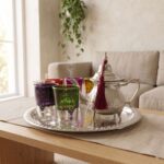 A steaming hand-engraved silver tea set multicolor glasses placed on a wooden coffee table with a linen runner, showcasing an ornate teapot and vibrant patterned glasses on a silver tray in a softly lit living room with a beige sofa and hanging green plant.