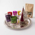 A traditional hand-engraved silver tea set multicolor glasses arranged on a matching silver tray, including an ornate teapot with a red tassel and six distinctively colored patterned glasses, displayed next to a kraft paper pouch of loose leaf green tea against a white background.