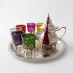A vibrant hand-engraved silver tea set multicolor glasses featuring a detailed teapot adorned with a red tassel and six brightly colored tea glasses with black geometric designs, all sitting on an intricately decorated silver tray against a clean white background.