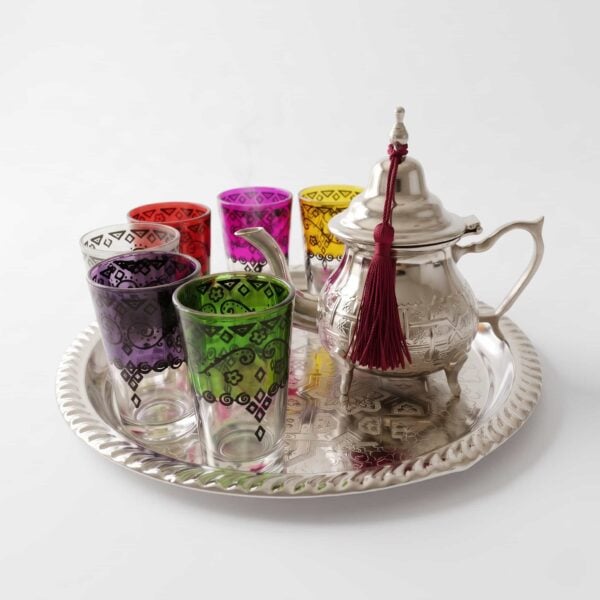 A vibrant hand-engraved silver tea set multicolor glasses featuring a detailed teapot adorned with a red tassel and six brightly colored tea glasses with black geometric designs, all sitting on an intricately decorated silver tray against a clean white background.
