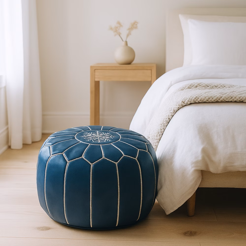 Minimalist bedroom scene featuring a leather pouf blue next to a light wood nightstand and white bedding.
