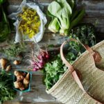 Natural beige straw basket bag with leather straps surrounded by garden vegetables and wildflowers