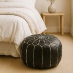 Round black leather floor ottoman pouf with white contrast stitching beside bed in minimalist bedroom