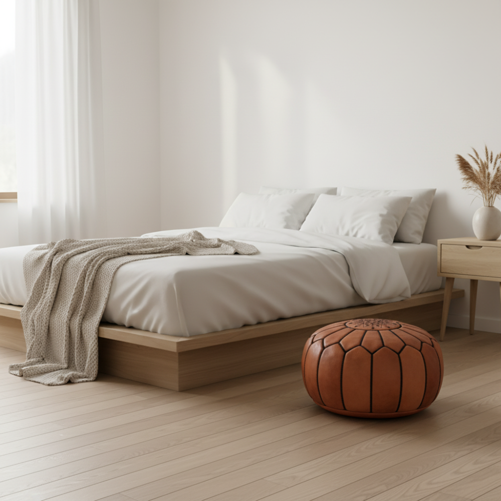 Natural leather pouf ottoman styled in a minimalist bedroom beside a bed with white linens.