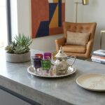 Silver tea set with multicolor glasses on a concrete counter near a succulent and leather lounge chair