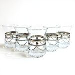 Studio lineup of six clear moroccan tea glasses with ornate silver chain bands, displayed against a white background.