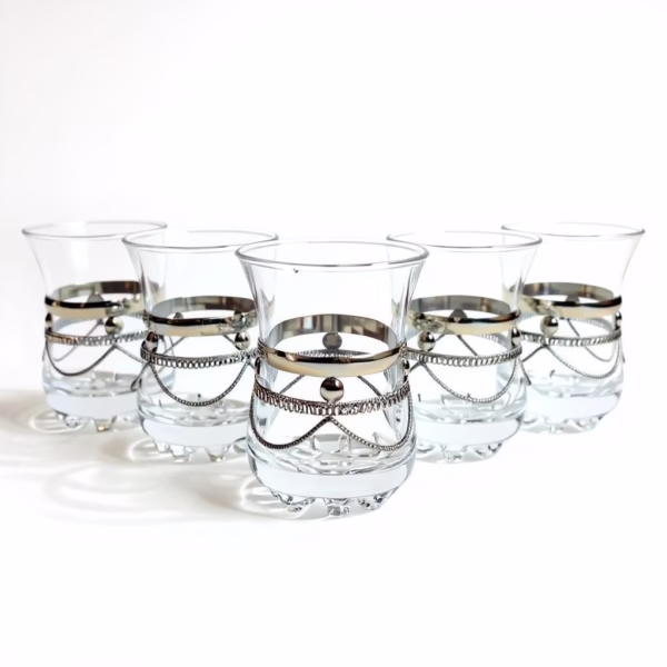Studio lineup of six clear moroccan tea glasses with ornate silver chain bands, displayed against a white background.