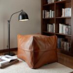 Light brown square leather ottoman as footrest beside bookshelf in reading nook, handmade Moroccan pouf