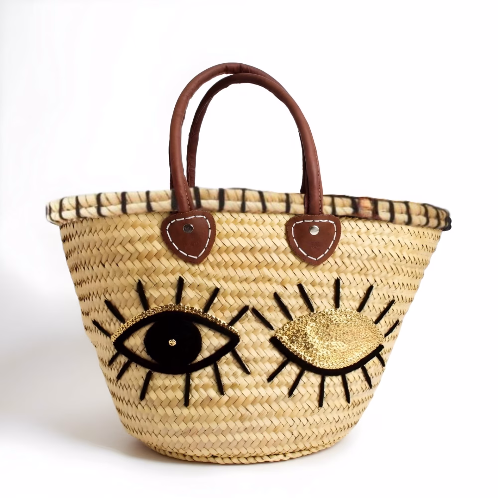 Handwoven straw tote bag with black and gold sequin winking eyes and leather handles on white background