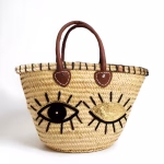 Handwoven straw tote bag with black and gold sequin winking eyes and leather handles on white background