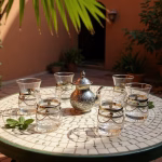 Sun-drenched courtyard scene featuring moroccan tea glasses and an engraved teapot on a mosaic patio table.