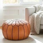 Tan leather round ottoman positioned next to a white armchair with a chunky knit throw blanket.