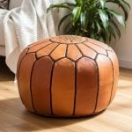 Leather round ottoman sitting on a wooden floor next to a large green houseplant and a cream sofa.