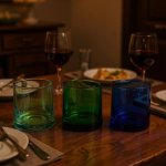 Trio of green, aqua, and cobalt moroccan wine glasses arranged on a wooden dinner table beside red wine goblets.