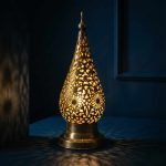 Illuminated Moroccan brass bedside lamp glowing against a dark blue background, casting intricate shadow patterns through its filigree design.
