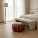 A brown leather round ottoman styled in a neutral bedroom setting, placed near the foot of the bed alongside a linen bench.