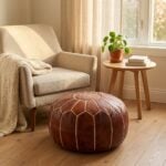 A brown leather round ottoman positioned as a footrest in a cozy corner next to a textured beige armchair and a wooden side table.