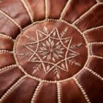 Close-up view of the intricate geometric embroidery and hand-stitching on the top center of a brown leather round ottoman.