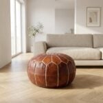 A brown leather round ottoman centered on a herringbone wood floor in a bright, minimalist living room with a modern beige sofa.