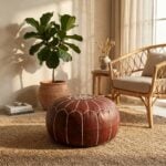 A brown leather round ottoman resting on a woven jute rug next to a rattan chair and a large potted plant in a warm, sunlit room.