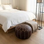 A dark brown leather round pouf placed at the foot of a bed next to a folded beige blanket and a black floor lamp in a cozy bedroom.