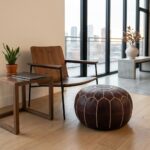A dark brown leather round pouf resting on a wooden floor next to a modern wood and metal accent chair with a city view in the background.