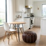 A dark brown leather round pouf styled on a light wood floor next to a small dining table in a bright, modern white kitchen.