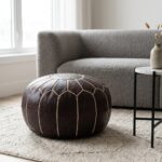 A dark brown leather round pouf sitting on a textured beige area rug in front of a contemporary gray boucle sofa and a marble side table.
